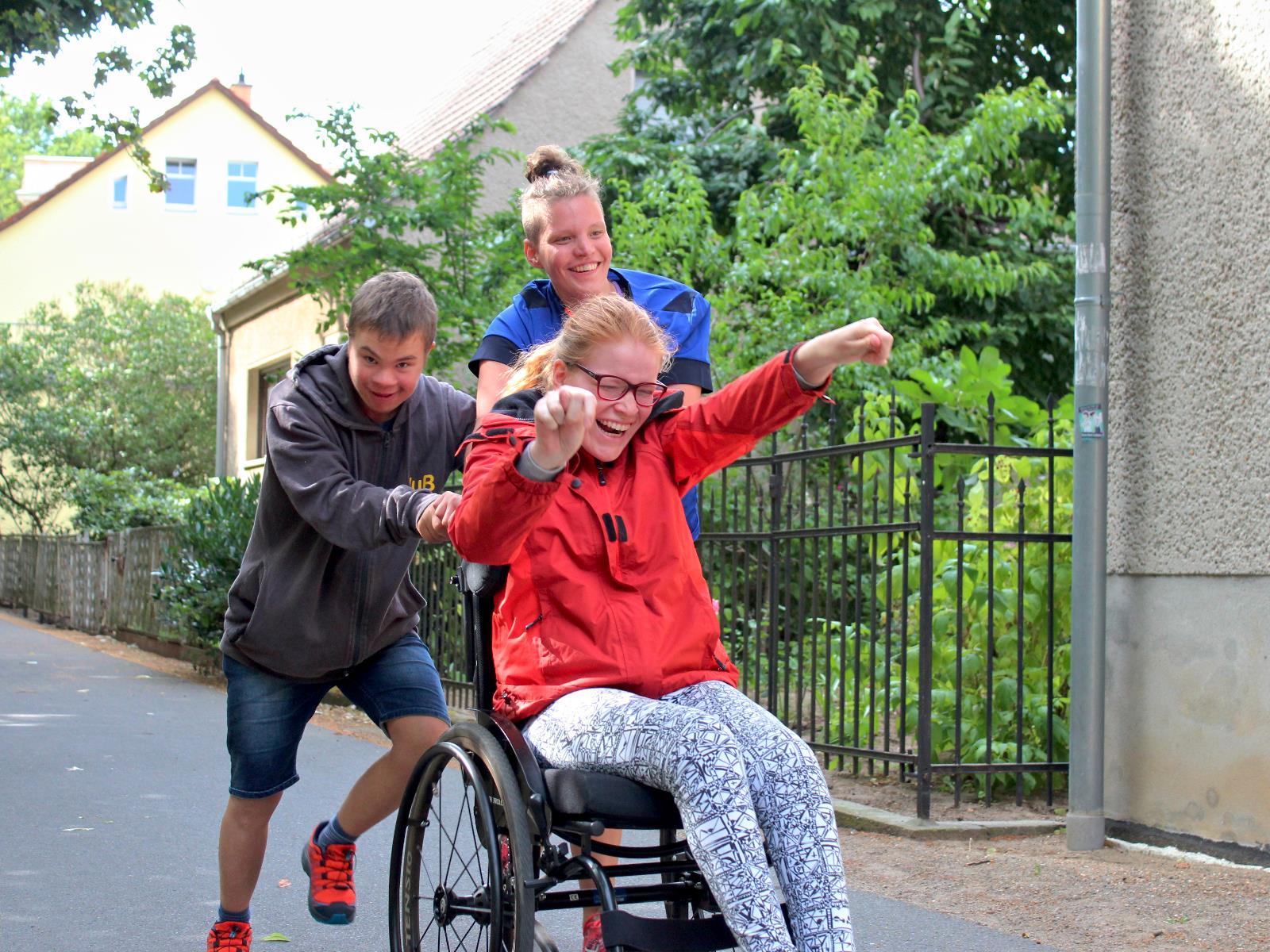 Zwei junge Menschen schieben Rollstuhlfahrerin. Alle haben Freude dabei.