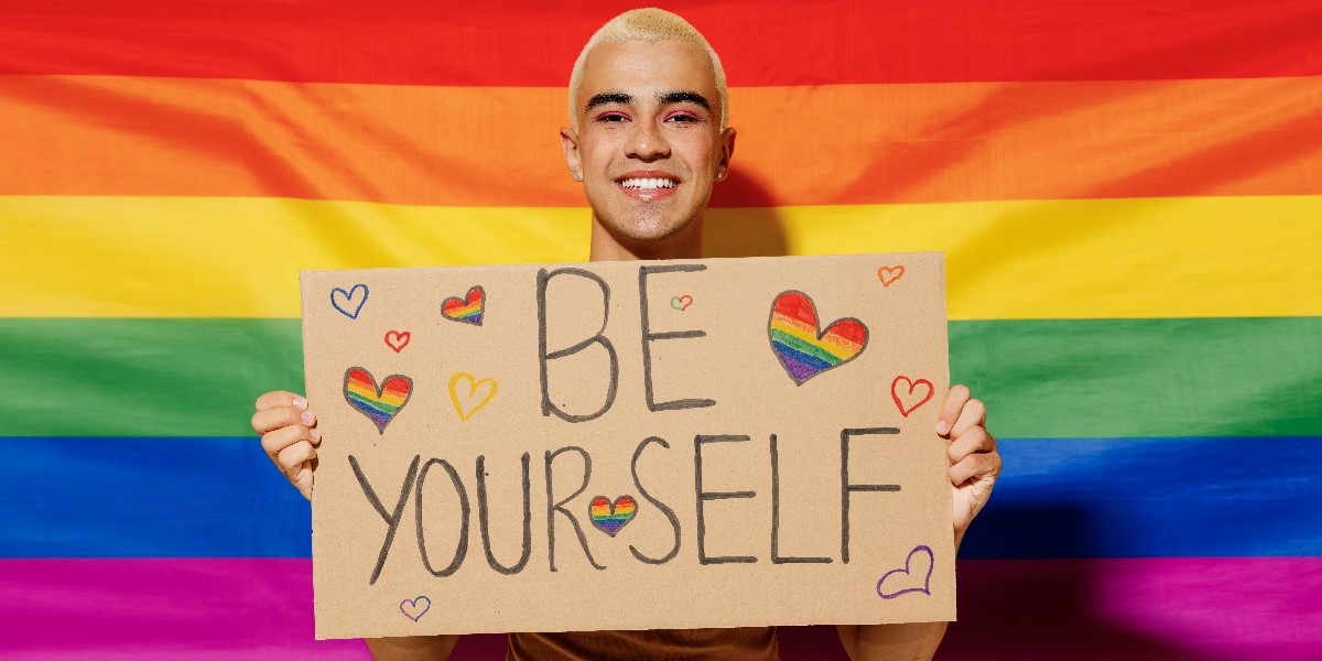 Queerer junger Mensch hält ein Plakat "Be yourself" und steht vor einer Regenbogenfahne