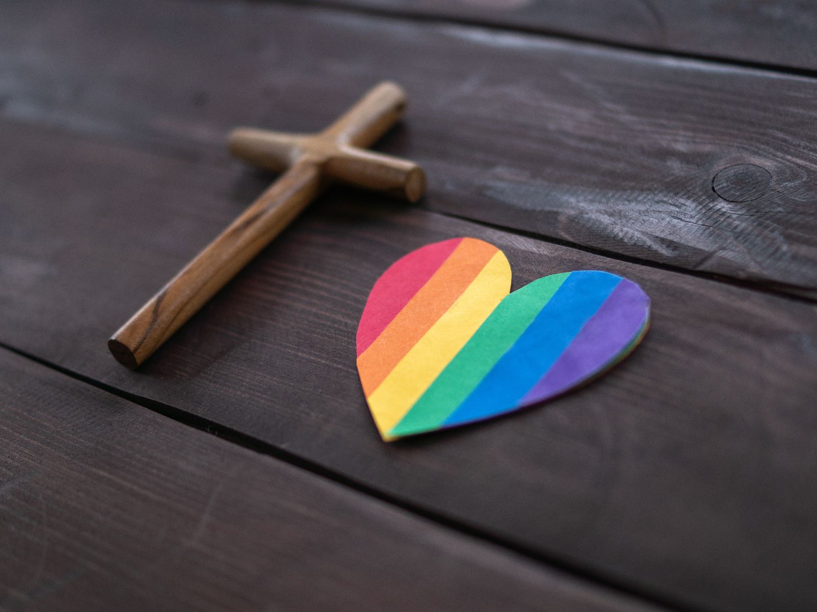 Holzkreuz und Herz in Regenbogenfarben liegen auf einem Holztisch.